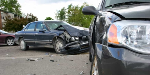 Close up of the front of a grey car that's been smashed in an accident, with another smashed blue car in the background
