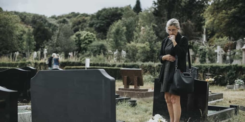 Older woman wearing black stands over a memorial plot in a cemetery with a sad look on her face