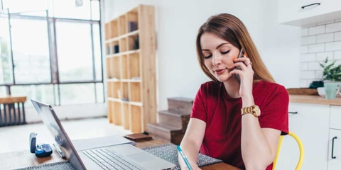 Woman with long brown hair sits at her kitchen table writing in a notebook with her laptop in front of her while she's on the phone