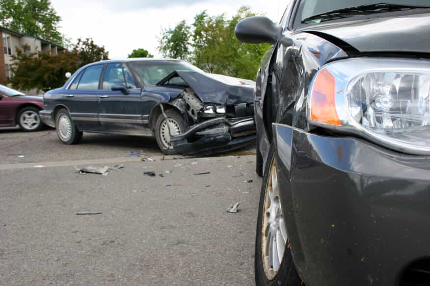 Close up of the front of a grey car that's been smashed in an accident, with another smashed blue car in the background