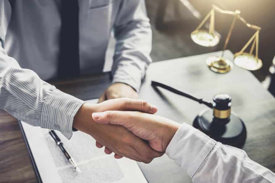 View from above of two men in long-sleeved button down shirts shaking hands over a table with a paper, pen, scales, and a gavel