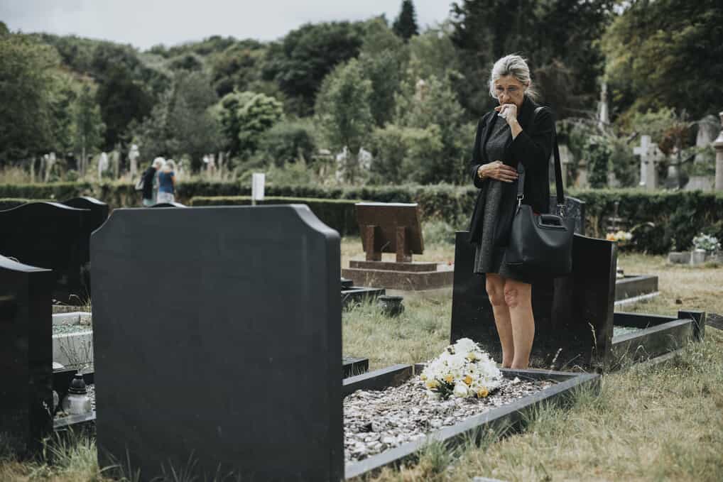 Older woman wearing black stands over a memorial plot in a cemetery with a sad look on her face