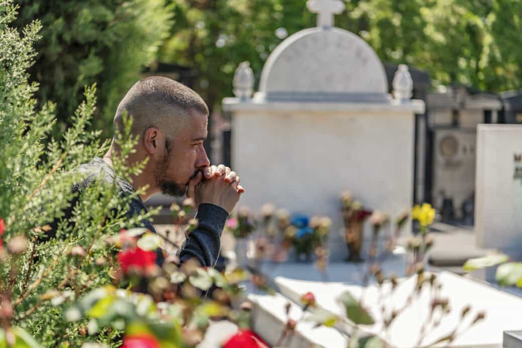 Man with military haircut sits with his hands at his face in sadness at a cemetery